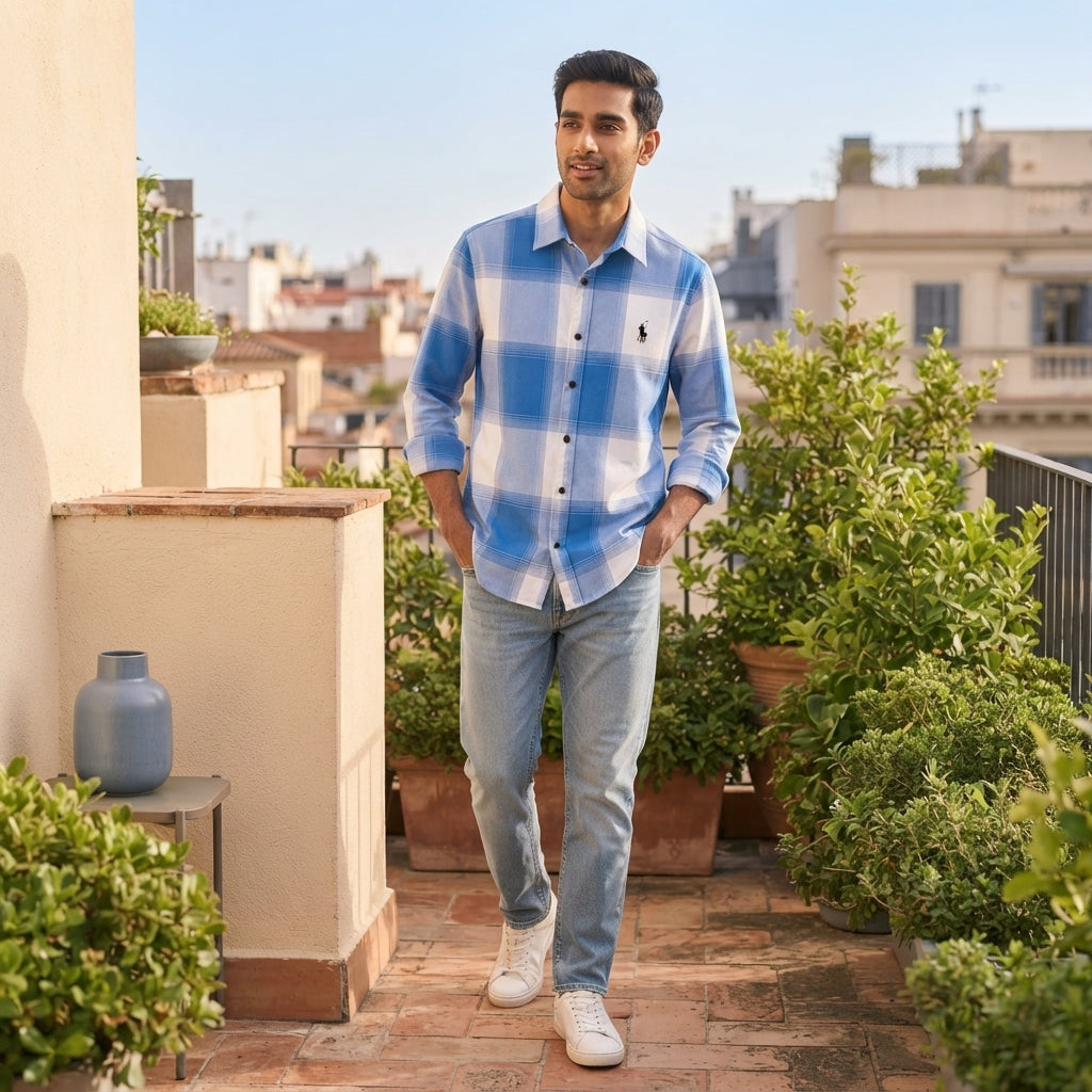 A South Asian man wearing a blue and white check shirt posing on a sunny balcony with cream and slate blue decor.