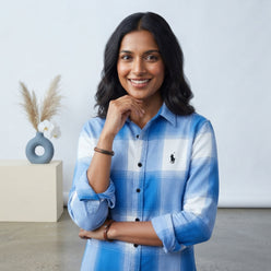 Anjali Singh posing in a blue and white check shirt against a clean, bright background for a professional editorial portrait.